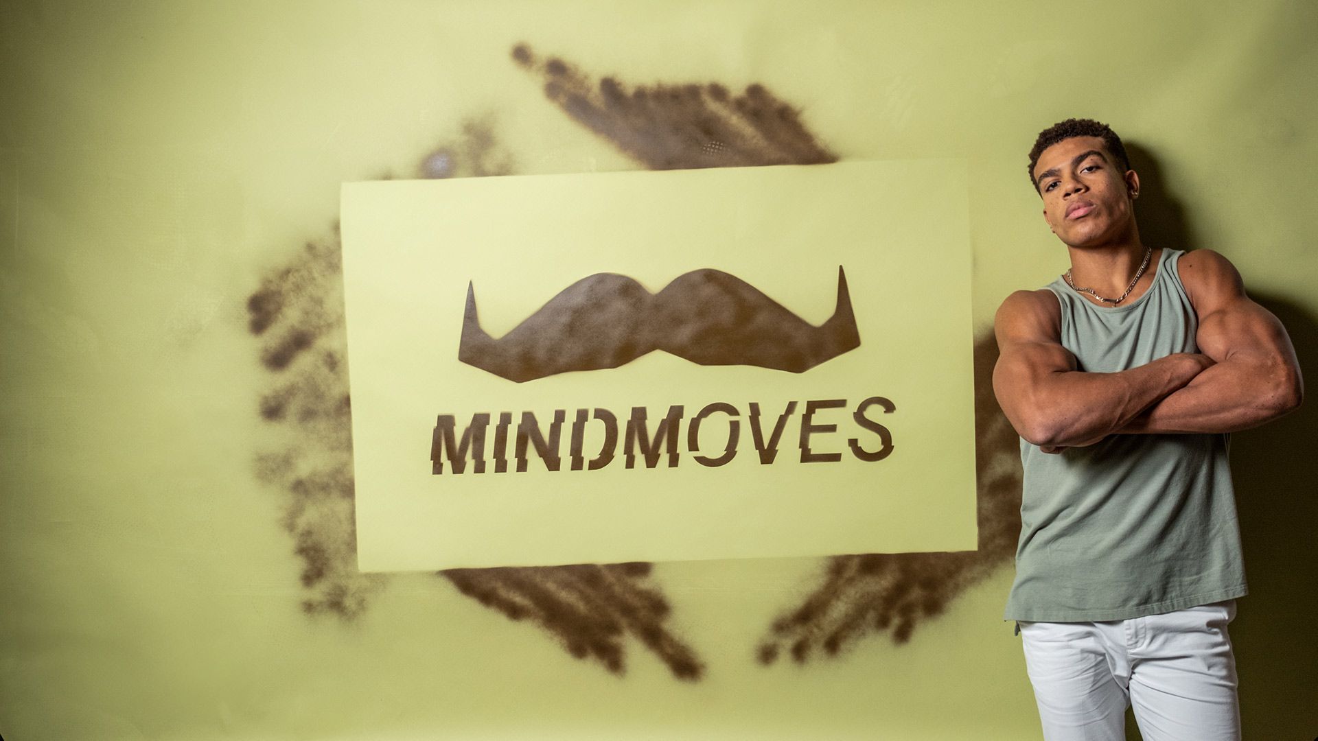 Photo of athlete Sam Phillips, posing to camera. A poster on the wall behind him says "MindMoves" above a furry-looking Movember moustache logo.