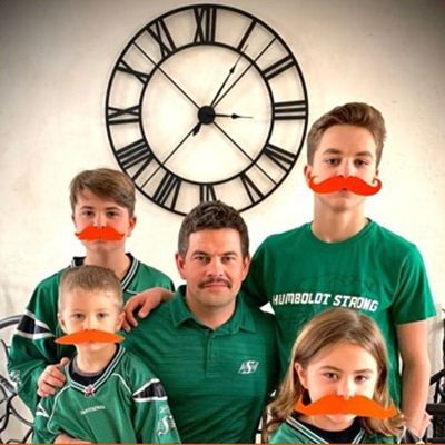 Man smiling to camera, flanked by his four children. All wear real or paper orange moustaches.