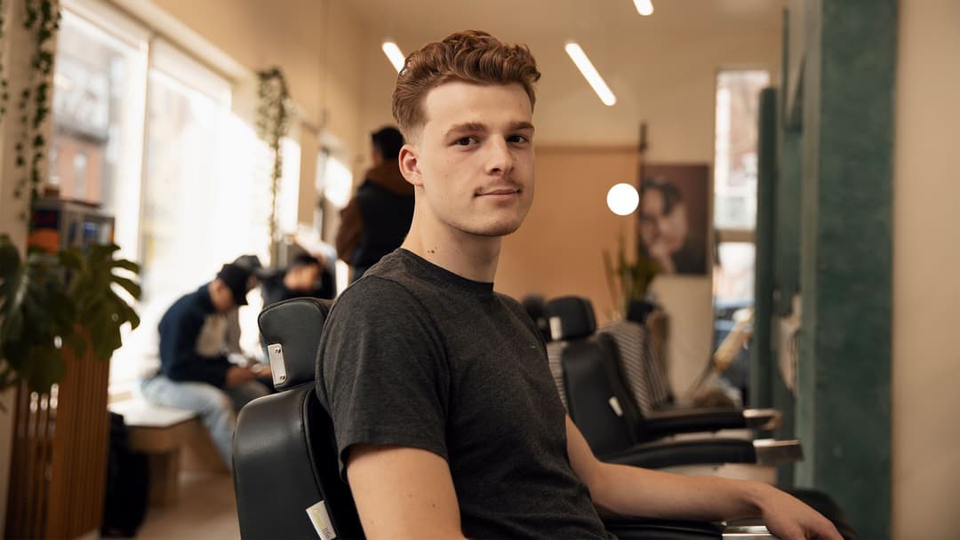 Un jeune étudiant sourit depuis le fauteuil de barbier d’un salon de coiffure montréalais