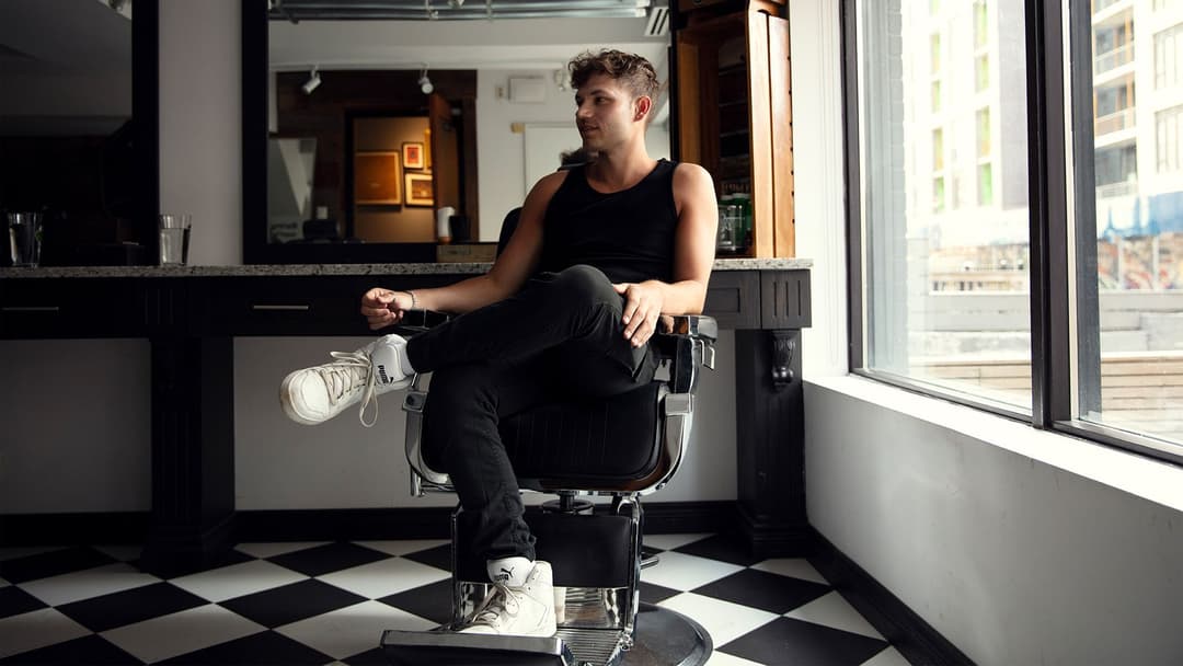 Un jeune homme est assis de façon décontractée dans un salon de barbier au plancher à carreaux noirs et blancs