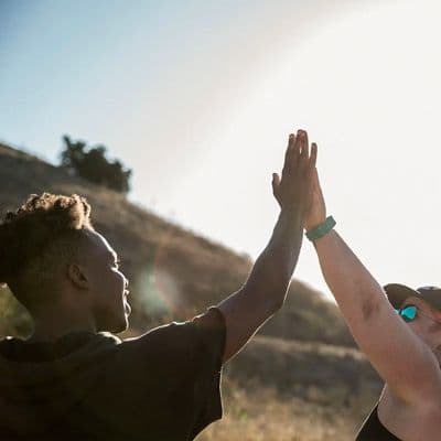 Two people high-fiving outdoors in bright sunlight against a hillside backdrop.