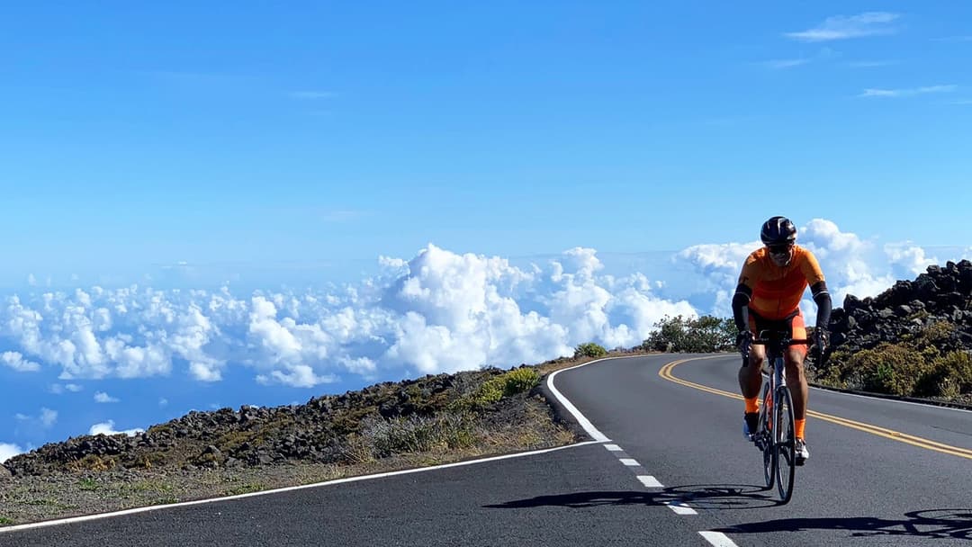 A man in an orange cycling suit cycles up a long road on Haleakalā Volcano in Maui. He's so high up that he's above the clouds.