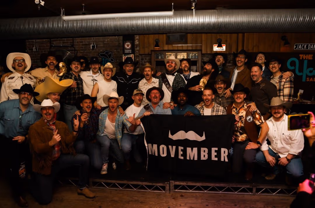 A team of Movember fundraisers posing in front of their camera with their mos and a Movember banner