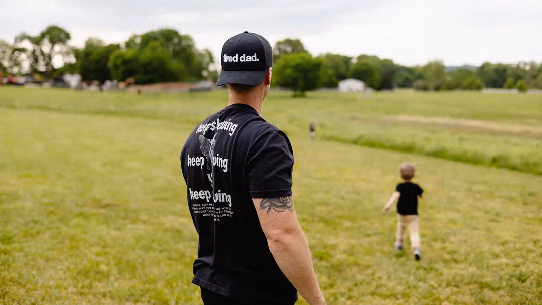 A man stands in a green field with his back to the camera, his young son runs ahead of him. The back of the man's hat reads "Tired Dad"
