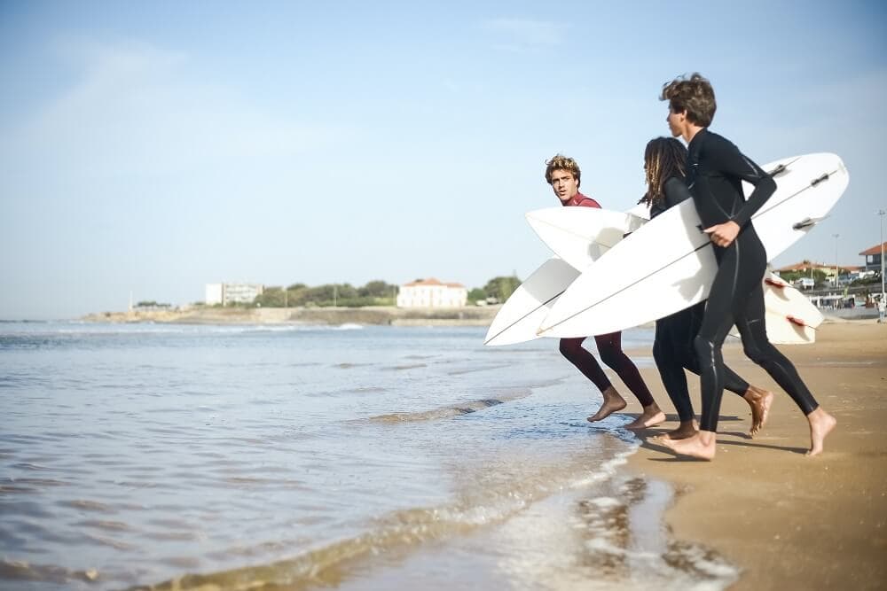 Three young men in wetsuits carrying surfboards along the shoreline.
