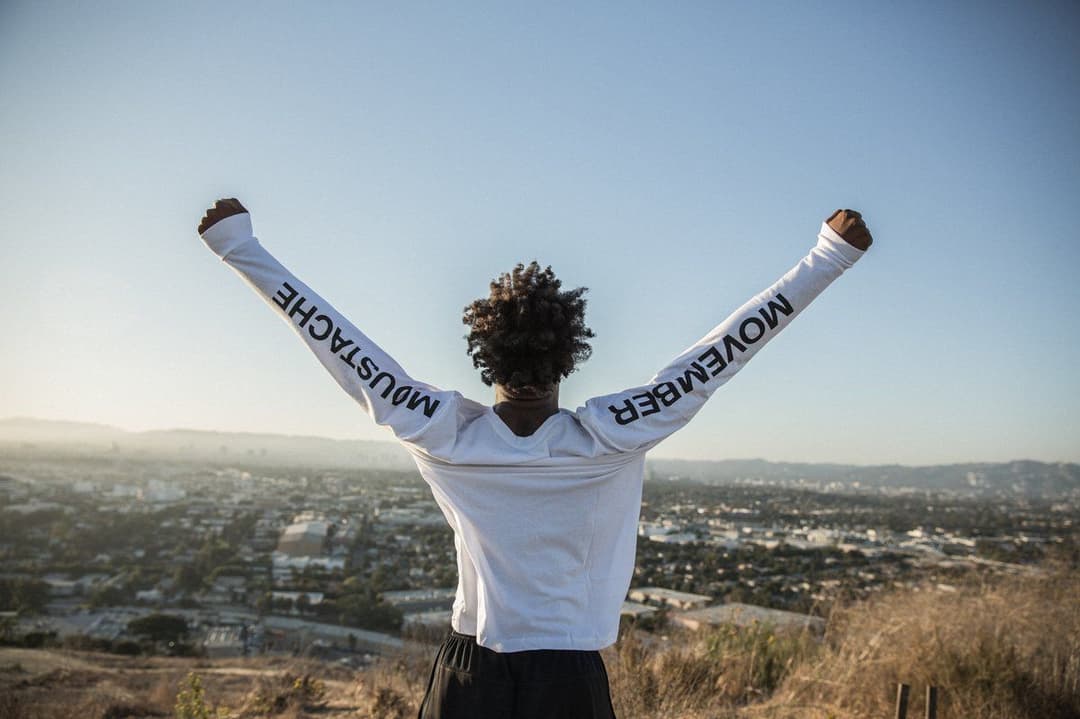 A man wearing a Movember long-sleeve shirt stands on a hilltop with arms raised, looking out over a city skyline.